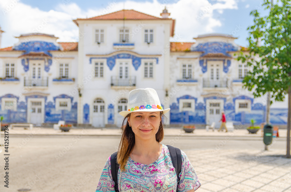 Young woman traveler with hat looking at camera posing and smiling in streets of Aveiro city in Portugal in sunny summer day with Aveiro station central railway azulejos-tiled building background