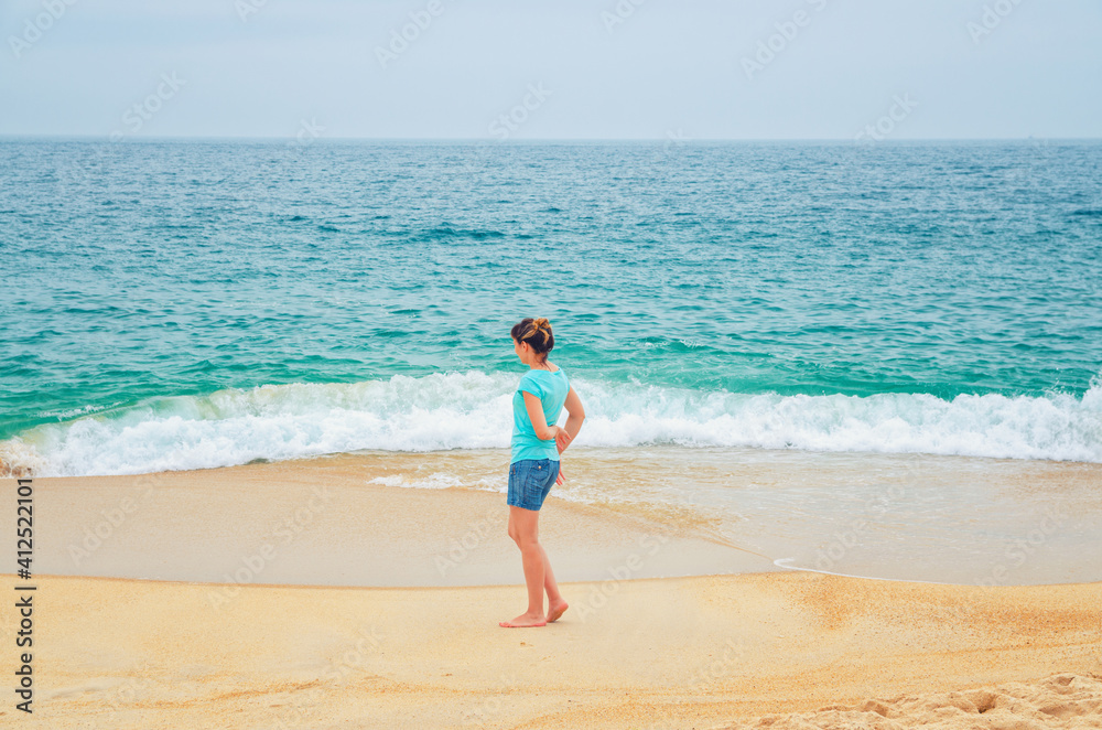 Young woman traveler walking down sandy beach coastline and looking at turquoise water, waves and endless horizon of Atlantic ocean, tourist female on vacation in Nazare town Portugal in summer day