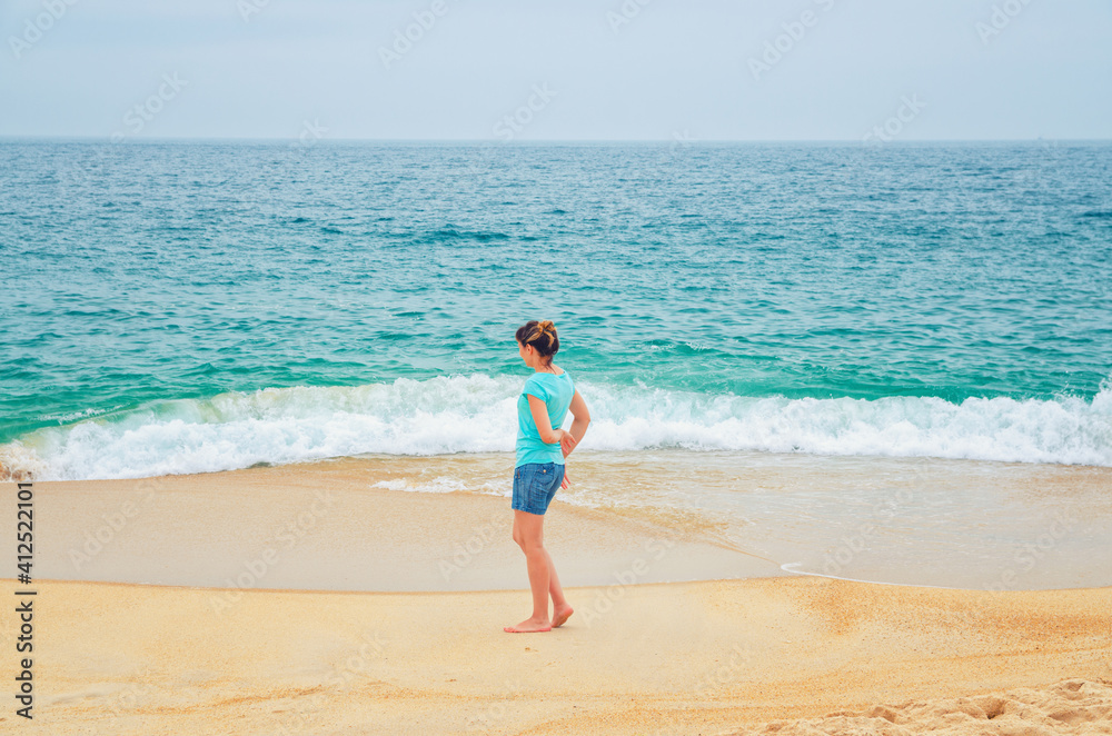 Young woman traveler walking down sandy beach coastline and looking at turquoise water, waves and endless horizon of Atlantic ocean, tourist female on vacation in Nazare town Portugal in summer day Young woman traveler walking down sandy beach coastline and looking at turquoise water, waves and endless horizon of Atlantic ocean, tourist female on vacation in Nazare town Portugal in summer day
