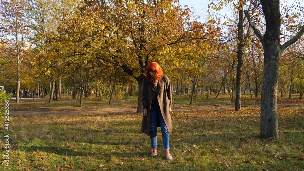 a girl with red hair in a coat, in an autumn park against the background of a yellow tree, is looking for something in the grass a girl with red hair in a coat, in an autumn park against the background of a yellow tree, is looking for something in the grass