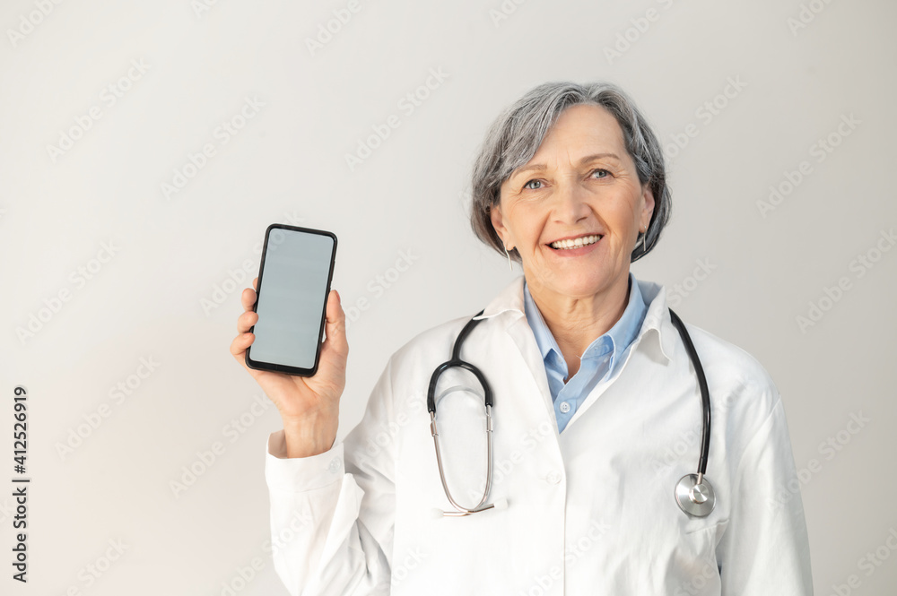 Senior mature female doctor with a stethoscope,posing isolated over gray background, demonstrating smartphone with a blank screen for advertisement, smiling healthcare professional showing digital app Senior mature female doctor with a stethoscope,posing isolated over gray background, demonstrating smartphone with a blank screen for advertisement, smiling healthcare professional showing digital app