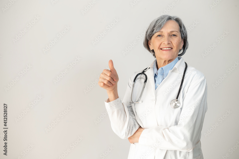 Positive senior mature female doctor with a stethoscope in a medical coat posing isolated over gray background, showing a thumb-up, smiling and welcoming healthcare worker looking at the camera Positive senior mature female doctor with a stethoscope in a medical coat posing isolated over gray background, showing a thumb-up, smiling and welcoming healthcare worker looking at the camera