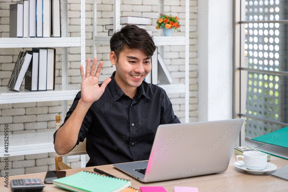 Young Asian businessman working at home with laptop in dining room, online meeting conference with colleague, and waving hand. Young Asian businessman working at home with laptop in dining room, online meeting conference with colleague, and waving hand.