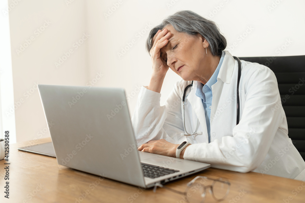 Tired stressed senior female doctor in a lab coat sitting in the office in front of a laptop, feeling burnout at work, restless after having no days off or exhausted, and falling asleep Tired stressed senior female doctor in a lab coat sitting in the office in front of a laptop, feeling burnout at work, restless after having no days off or exhausted, and falling asleep