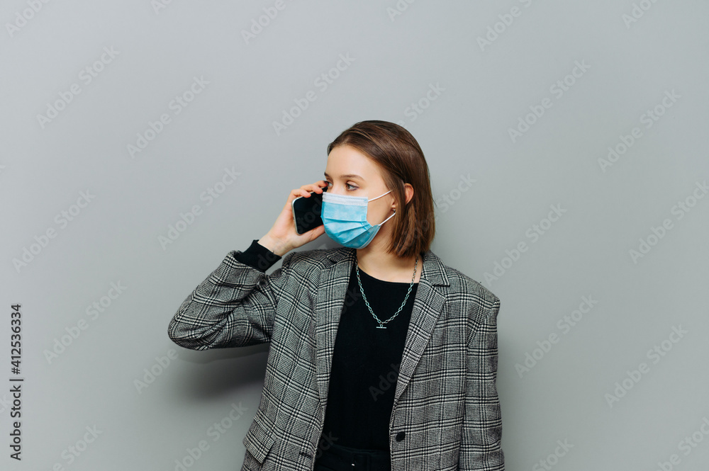 Beautiful business woman in formalwear and a medical mask on her face communicates on the phone on a background of a gray wall and looks away at an empty place, isolated. Beautiful business woman in formalwear and a medical mask on her face communicates on the phone on a background of a gray wall and looks away at an empty place, isolated.
