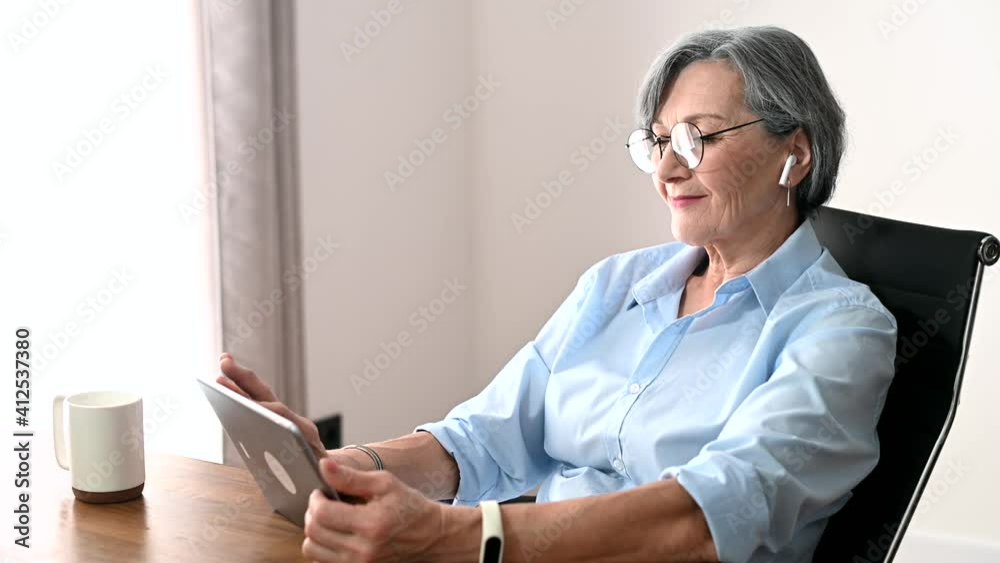 Charming senior businesswoman in glasses sitting at the desk, using a tablet for video chatting, an elder female English teacher is scrolling news online, looking at the photos and smiling Charming senior businesswoman in glasses sitting at the desk, using a tablet for video chatting, an elder female English teacher is scrolling news online, looking at the photos and smiling