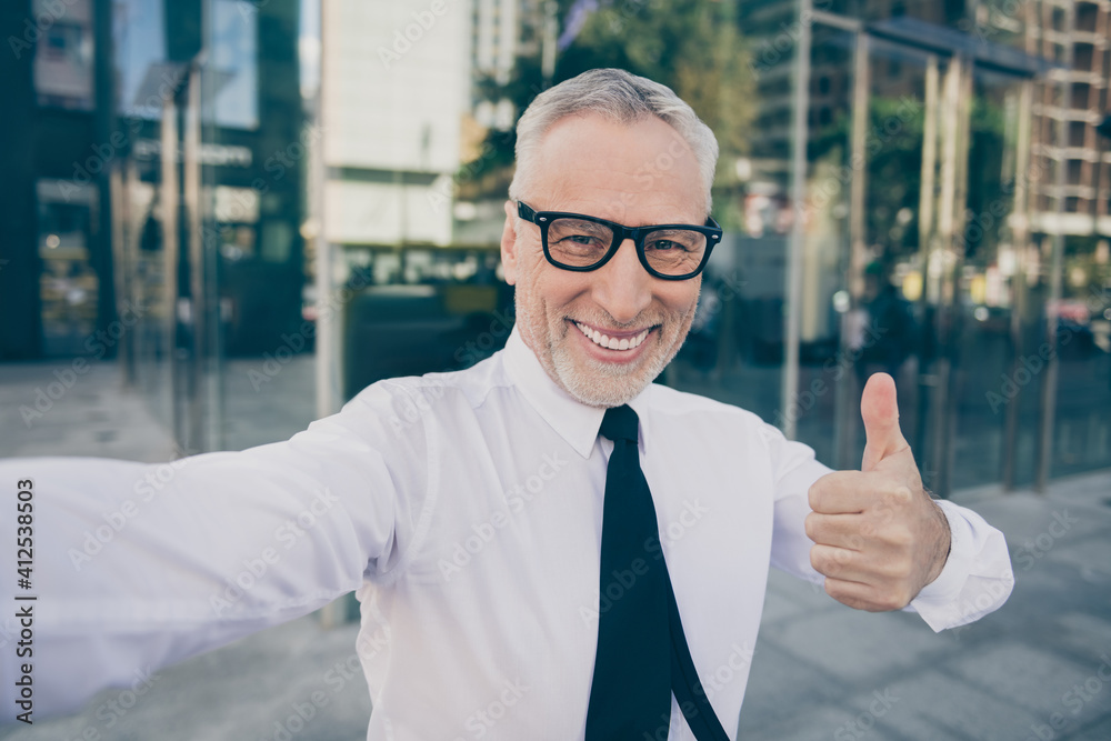 Photo of optimistic grey hair old business man show thumb up do selfie wear spectacles white shirt outdoors near work center Photo of optimistic grey hair old business man show thumb up do selfie wear spectacles white shirt outdoors near work center