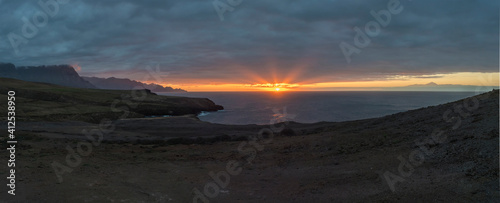 Panorama of beautiful sunset on the rocky atlantic coast in north west part of Gran Canaria island. Sun with rays going down to the ocean. Cliffs and Pico del Teide volacano in background.