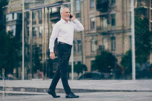 Photo of cute attractive mature worker man wear formal shirt walking talking modern device outdoors city street