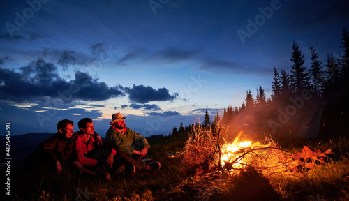 Summer recreation three tourists in the mountain camping with bonfire. Happy guys funny watching how burning dry brushwood on the backgraund of dusk sky.