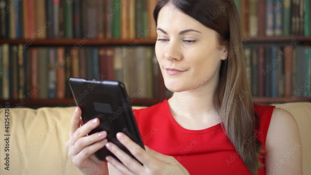 Young businesswoman at home reading e-book during lockdown. Bookshelves with books in the background. Paper vs digital reading, e-books replacing physical print books. Concept of social distancing