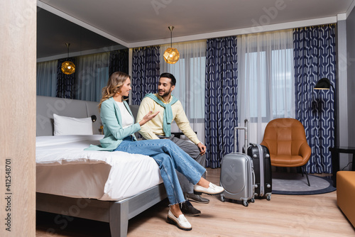 Smiling woman pointing with hand near suitcases and muslim boyfriend on bed in hotel