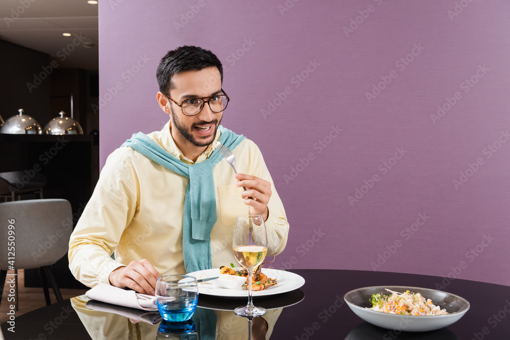 Smiling arabian man holding fork near meal and wine in hotel room Smiling arabian man holding fork near meal and wine in hotel room