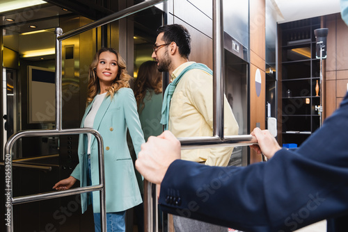 Smiling woman looking at muslim boyfriend near elevator and hotel porter on blurred foreground