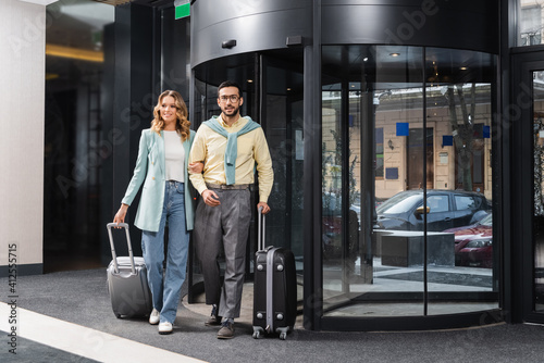 Smiling interracial couple walking with suitcases near hotel