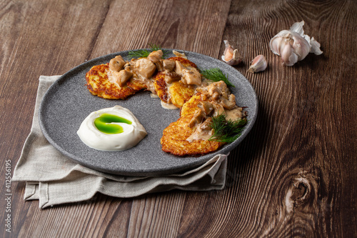 Fried potato pancakes served with mushrooms, sour cream and dill on a grey round plate. Close-up isolated on wooden background with grey linen napkin, sliced bread and garlic.