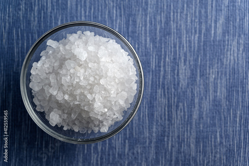 Glass bowl of sea salt on blue background with empty space. Shallow depth of field