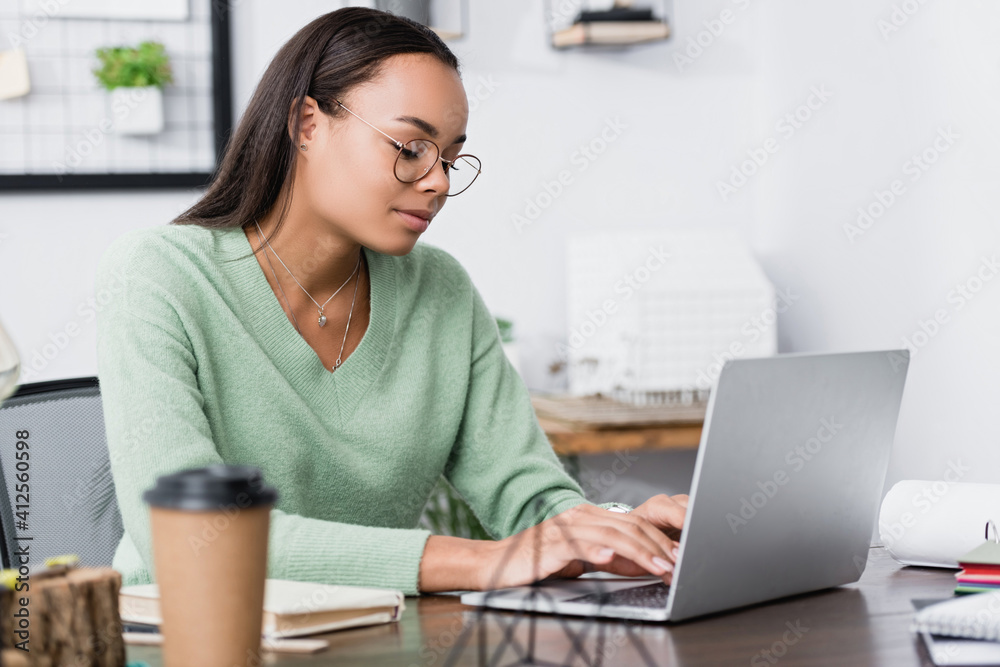 young african american architect typing on laptop near cup on blurred foreground young african american architect typing on laptop near cup on blurred foreground