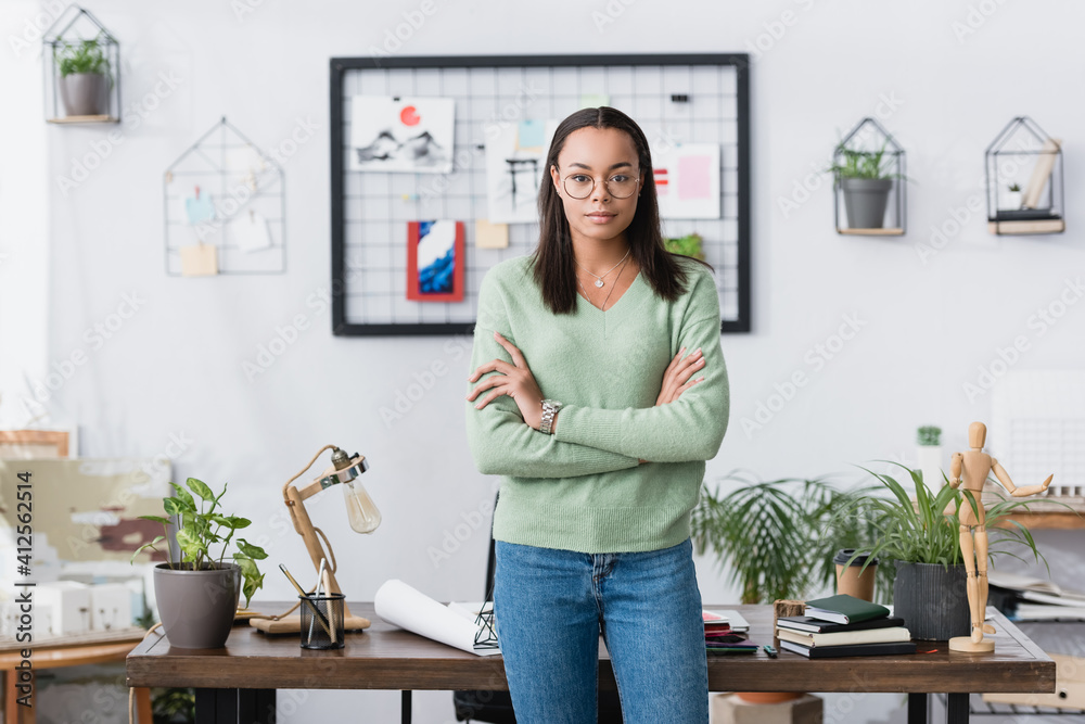 confident african american architect standing near workplace with crossed arms and looking at camera confident african american architect standing near workplace with crossed arms and looking at camera