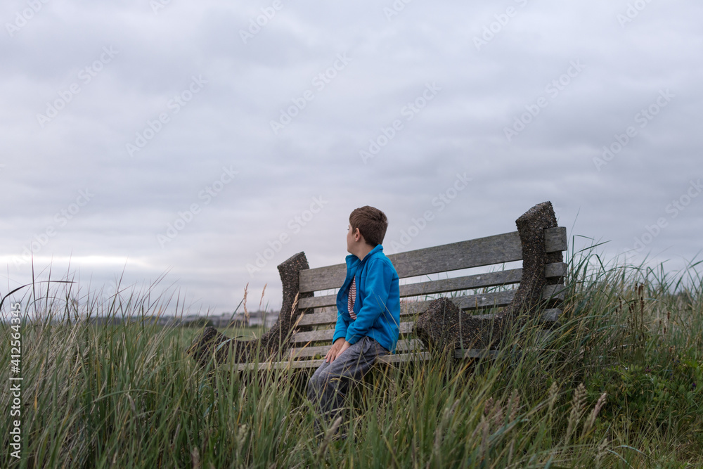 Young boy in blue fleece coat sits on wood and concrete bench in a field to beach grass; he looks away from camera out towards the horizon Young boy in blue fleece coat sits on wood and concrete bench in a field to beach grass; he looks away from camera out towards the horizon
