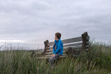 Young boy in blue fleece coat sits on wood and concrete bench in a field to beach grass; he looks away from camera out towards the horizon