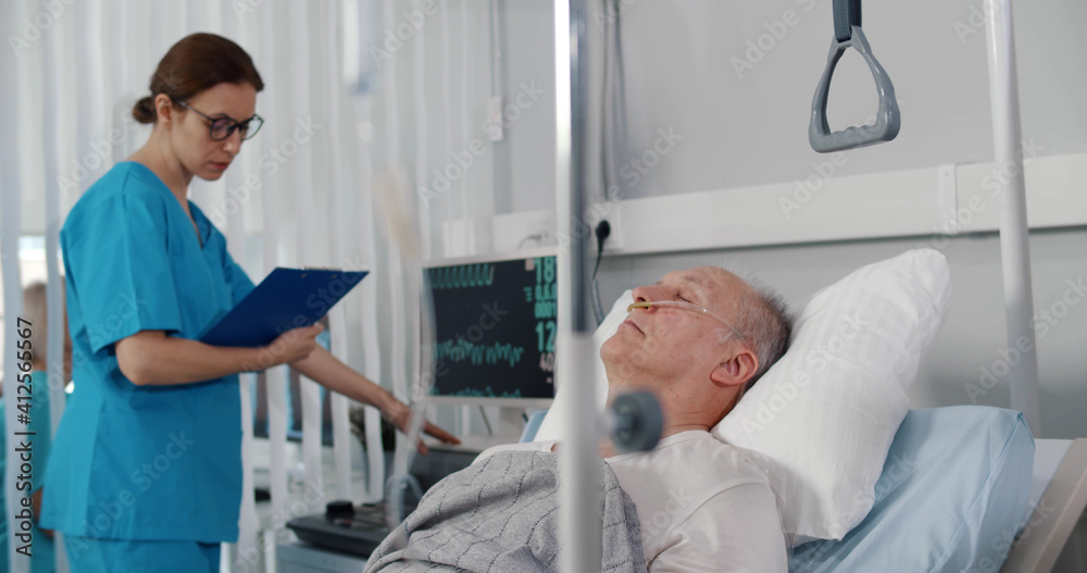 Obraz premium Doctor checking data on medical monitor while senior aged patient sleeping in bed at hospital ward