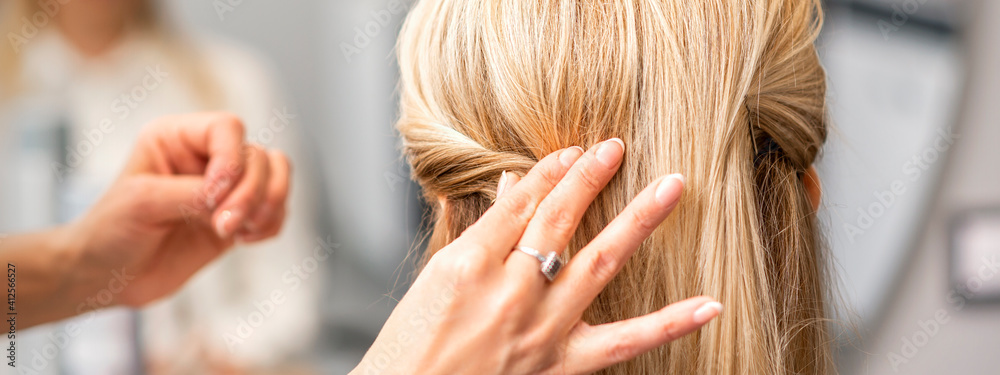 Fototapeta premium Back view of the female hand of hairdresser models a hairstyle of a young blonde woman in a hair salon