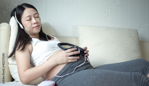Relaxation in pregnancy. Calm pregnant young asian girl Listen to music Mozart effect good for fetus by using headphones attached to the stomach hold earphones near belly lying on sofa at home.