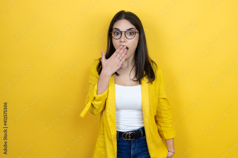 Young brunette businesswoman wearing yellow blazer over yellow background covering mouth with hand, shocked and afraid for mistake. surprised expression Young brunette businesswoman wearing yellow blazer over yellow background covering mouth with hand, shocked and afraid for mistake. surprised expression