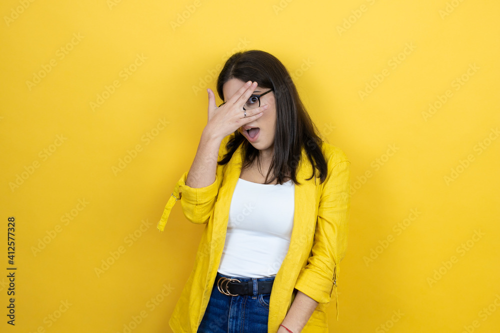 Young brunette businesswoman wearing yellow blazer over yellow background peeking in shock covering face and eyes with hand, looking through fingers with embarrassed expression Young brunette businesswoman wearing yellow blazer over yellow background peeking in shock covering face and eyes with hand, looking through fingers with embarrassed expression