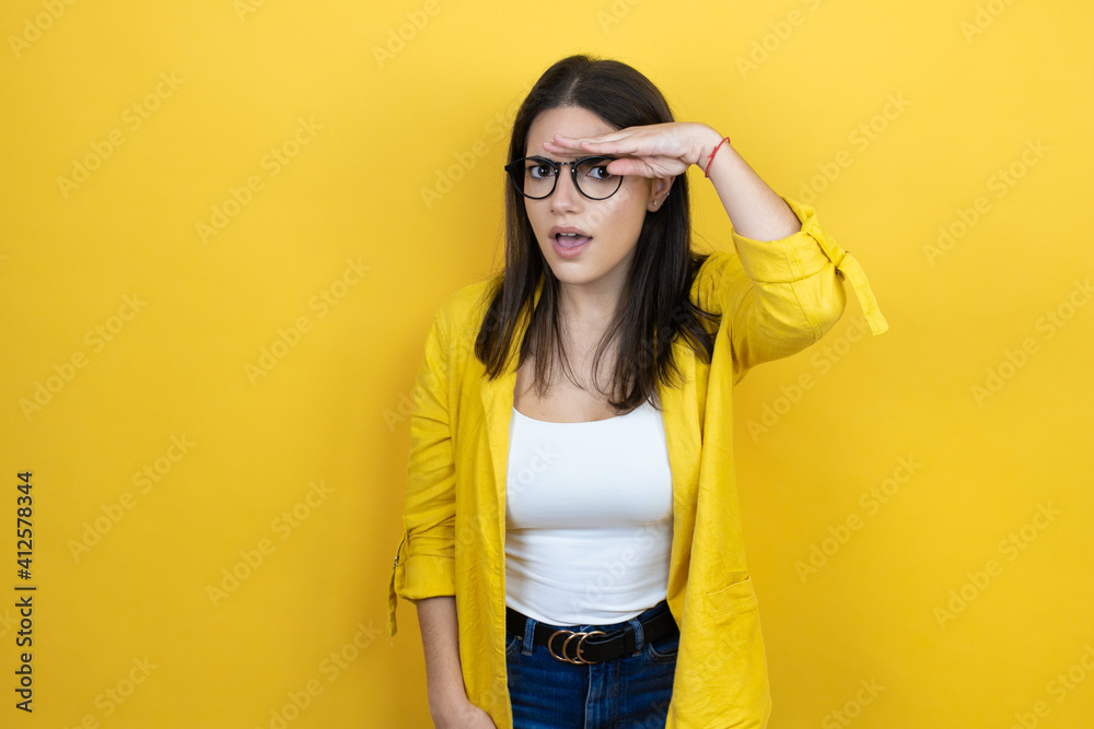 Fototapeta premium Young brunette businesswoman wearing yellow blazer over yellow background very happy and smiling looking far away with hand over head. searching concept.