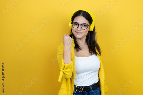 Young businesswoman wearing yellow blazer listening to music using headphones over yellow background very happy and excited making winner gesture with raised arms, smiling and screaming for success