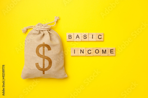 Basic income written on wood blocks with bag with money on yellow background. Back to basics fundamental principles concept