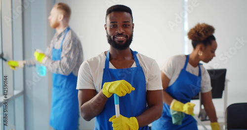 Portrait of african cleaner in apron and gloves holding mop and smiling at camera working in office photo