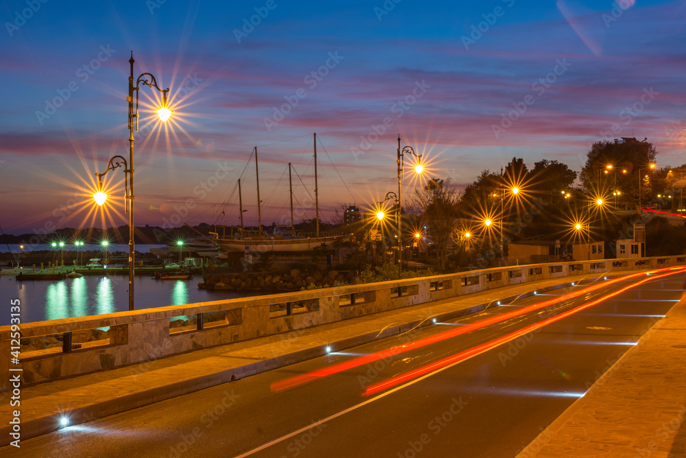 Colorful sunset and light trails motion of the passing by cars. Long exposure view of a harbour, near at the entrance of the UNESCO – protected ancient town of Nessebar, Bulgaria. Tourism concept.