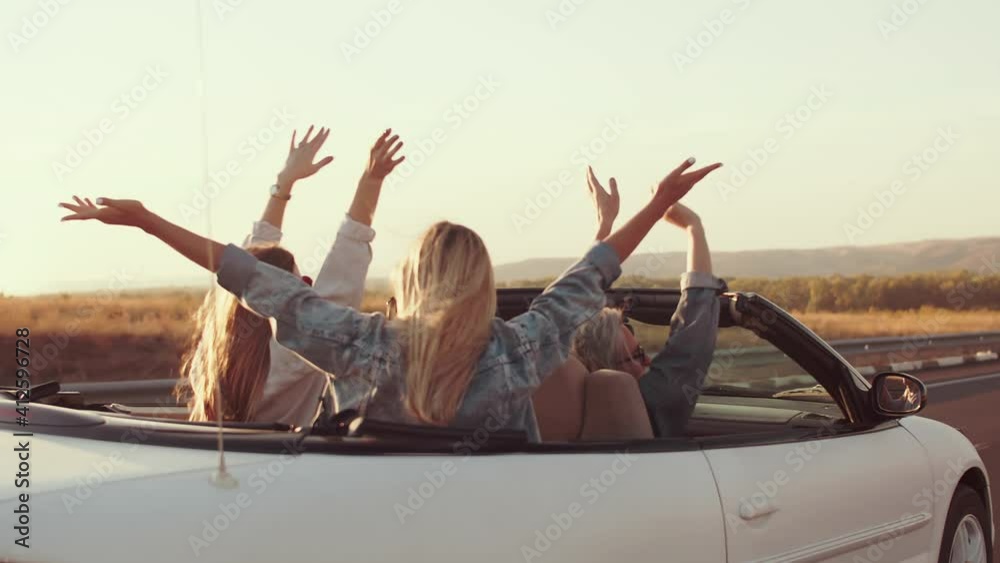 Group of friends ride in cabriolet car waving their hands from side to side, enjoying life, rear view. Friends in cabriolet car have fun wanderlust