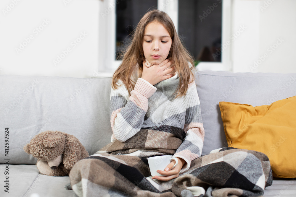 Pharyngitis, sore throat concept. Sick Caucaisna teenage girl touching sore throat, feeling unwell, while sitting on the sofa at home, covered with blanket and drinking hot tea