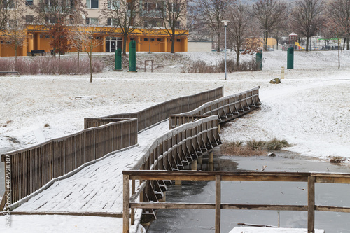 Wooden bridge over a pond in a city park. There is snow on the roads and meadow.