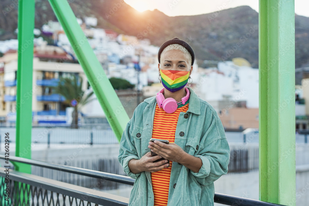 Obraz premium Beautiful short hair woman holing smartphone while wearing lgbt rainbow face mask for coronavirus outbreak