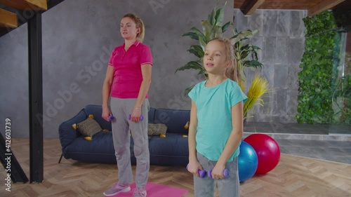 Sporty fit mother with cute preadolescent girl in activewear performing dumbbell upright row exercise at home, flexing front and middle deltoids, trapezius, rhomboids muscles during strength training photo