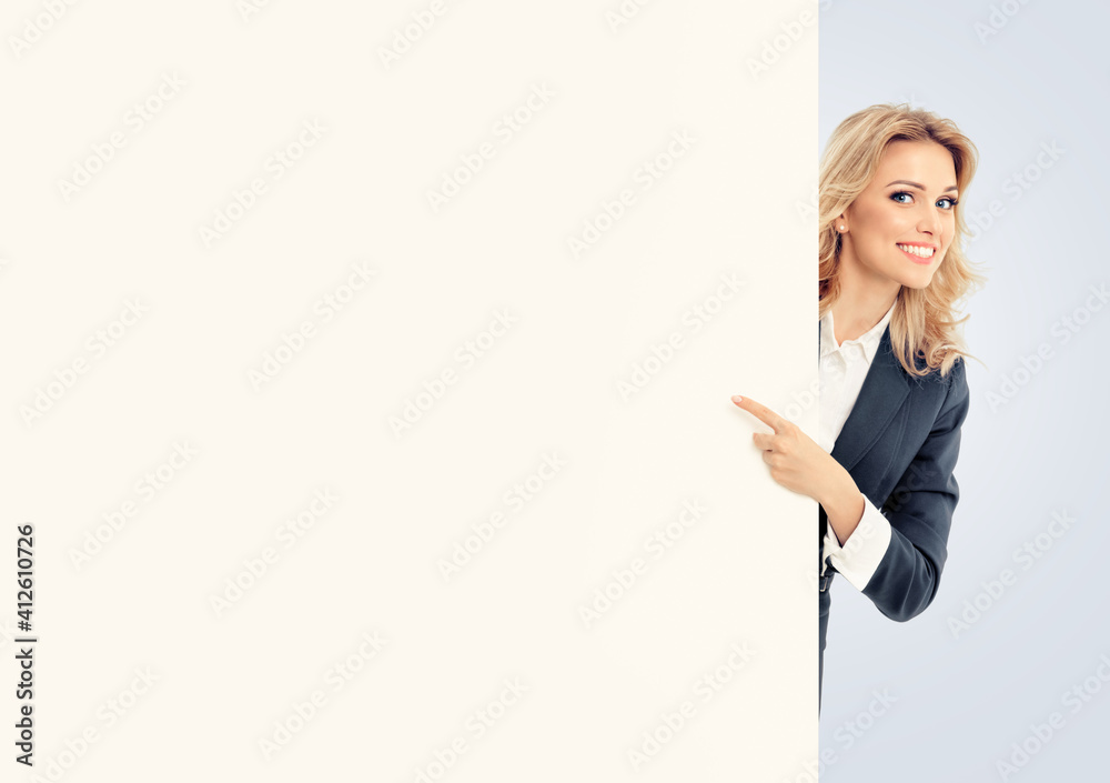 Happy smiling pointing businesswoman in grey confident suit, standing behind blank banner or signboard  with copy space for some text, isolated over gray background.