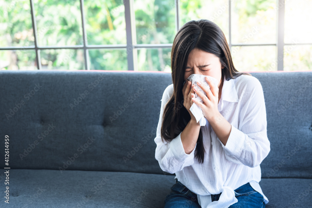 Portrait images of Asian attractive woman sitting on the sofa at home, is using tissue paper to wipe his nose because of coughing and sneezing due to flu, to people health care and virus infection Portrait images of Asian attractive woman sitting on the sofa at home, is using tissue paper to wipe his nose because of coughing and sneezing due to flu, to people health care and virus infection