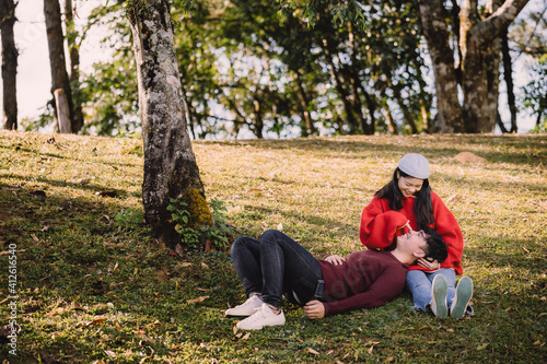 Portrait sweet couple of teenage sitting on hill, asian man with maroon sweater lay down on lap  young woman with red sweater. Romantic mood and sweet emotional relationship in summer with copy space