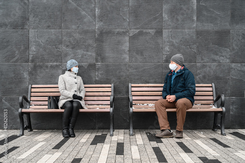Young caucasian man and woman keeping social distance and wearing protective face masks, chat together looking at each other. They sit on benches in the city, wintertime. Concept new normality