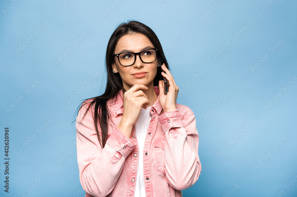 Pensive beautiful caucasian brunette woman talking on mobile phone with friends, family or boyfriend, smiling, looks away thoughtfully, standing against isolated blue background.