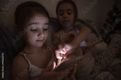 Two little girls sit in bed at night and use smartphones.