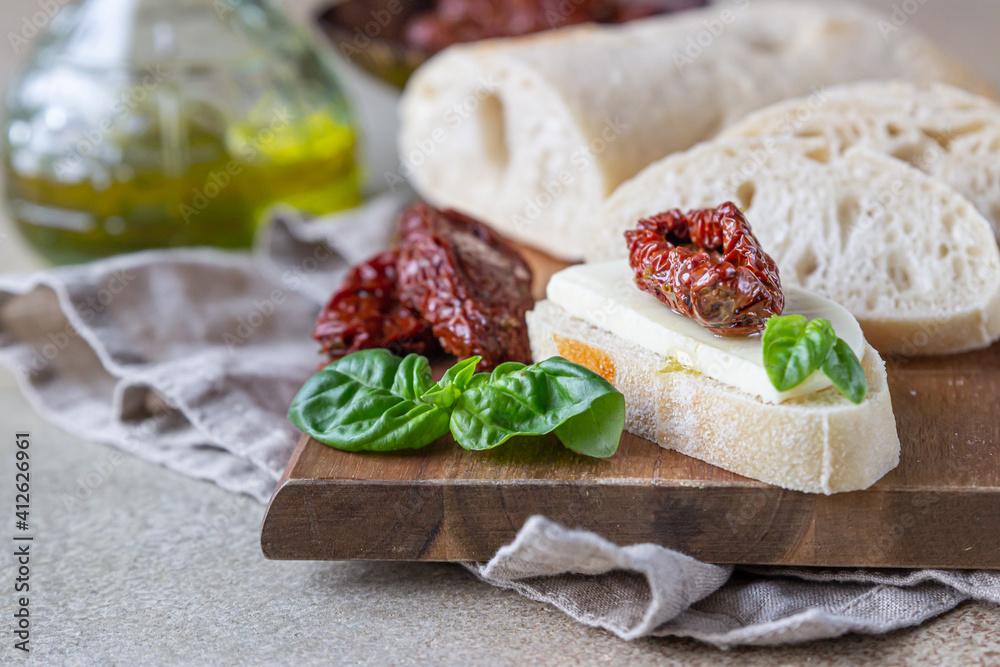 Sliced ciabatta bread with sun-dried tomatoes, cheese, basil and olive oil on wooden board, stone background.