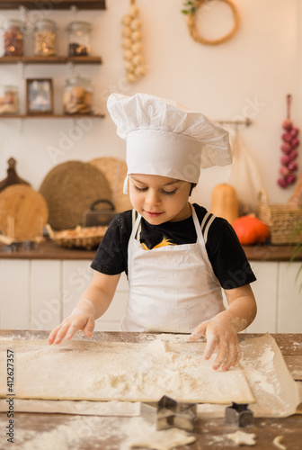 little chef in a white apron and cap kneads dough with his hands in the kitchen