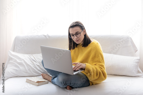 A cute girl in a yellow sweater and glasses works at a laptop at home on the couch.