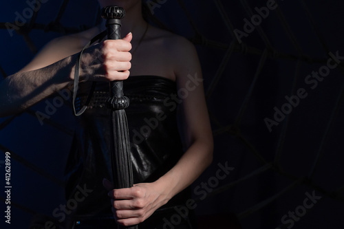 BDSM concept. A woman in a leather corset holds a black lash with a long fringe. Role-playing games for adults. Close-up of sex toys for domination. The whip in female hands.